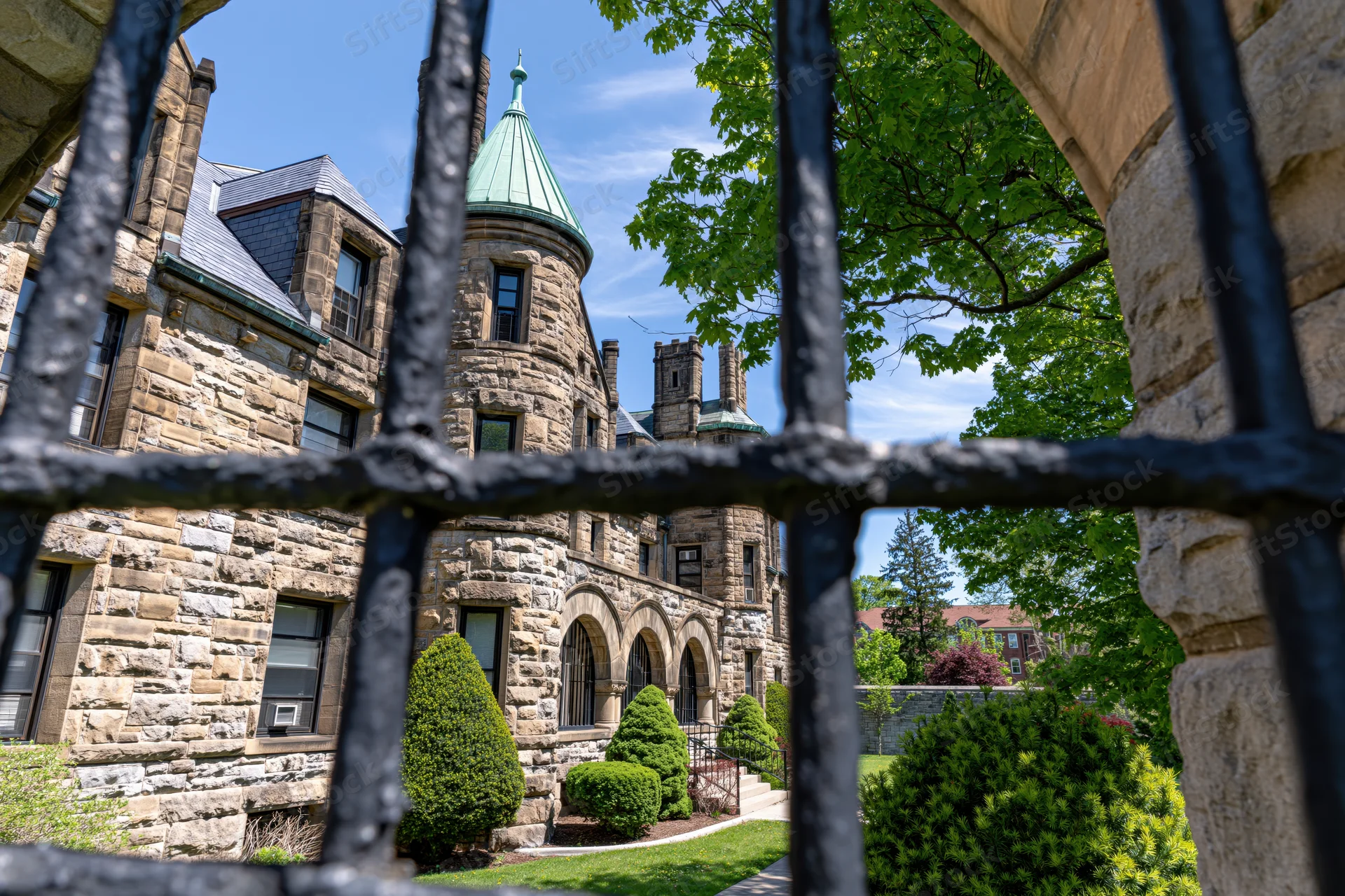 Historic stone prison exterior viewed through iron gate with greenery and turret under blue sky