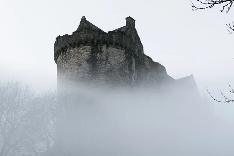 Historic stone castle with round crenellated tower emerging from fog among bare trees
