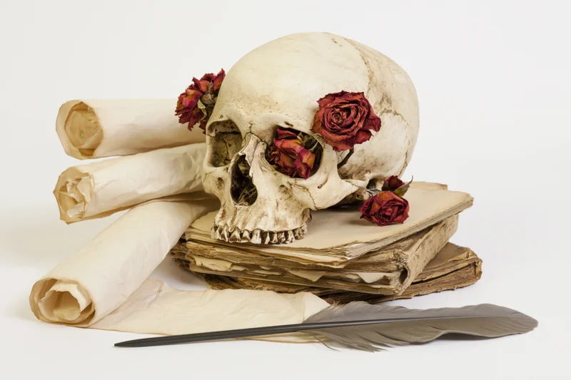 Human skull adorned with dried roses resting on a stack of antique books and scrolls with a feather quill in front