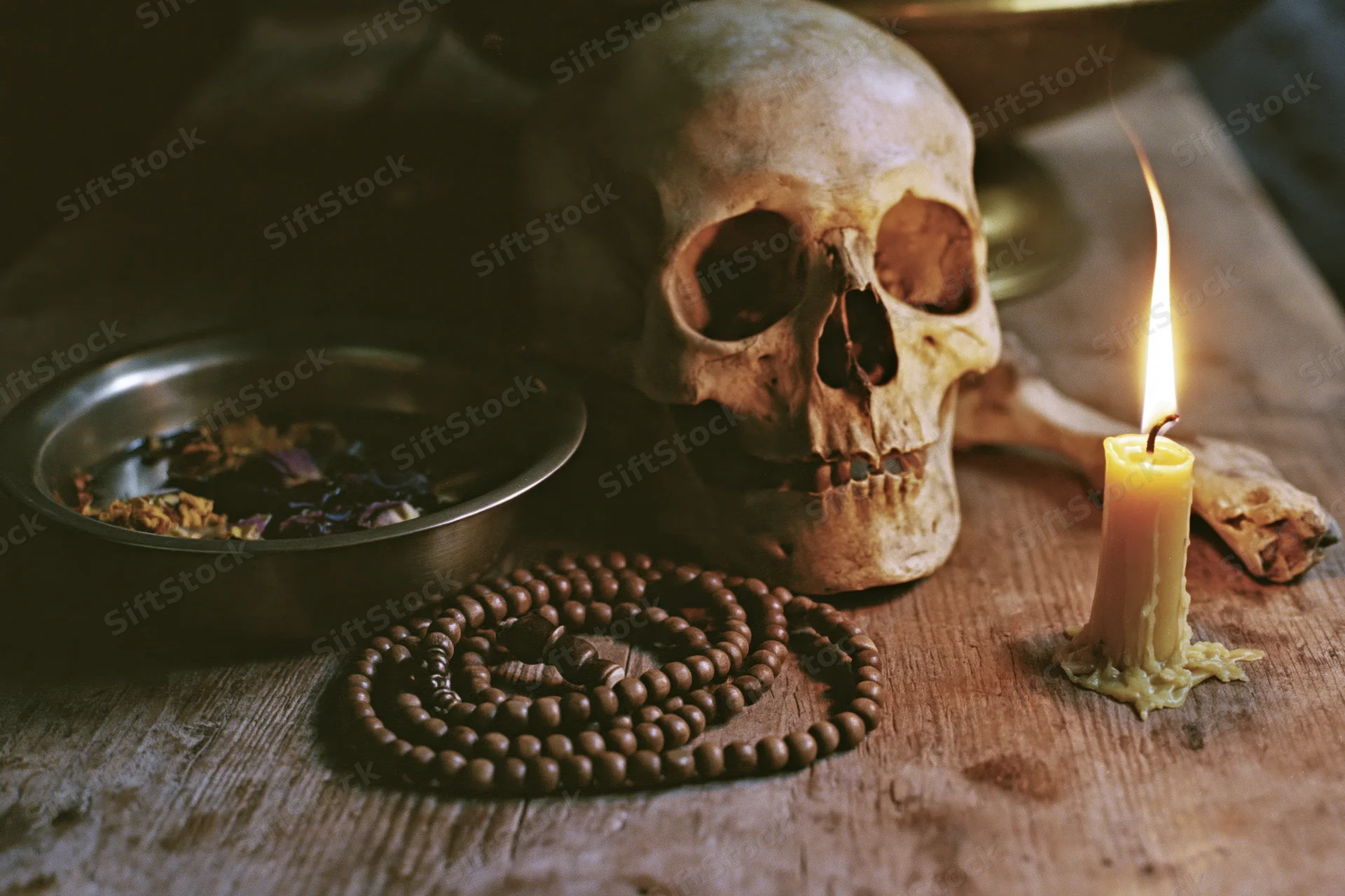 Human skull on wooden table with lit candle, beads, bowl of herbs, and bone fragment, dimly lit by flame