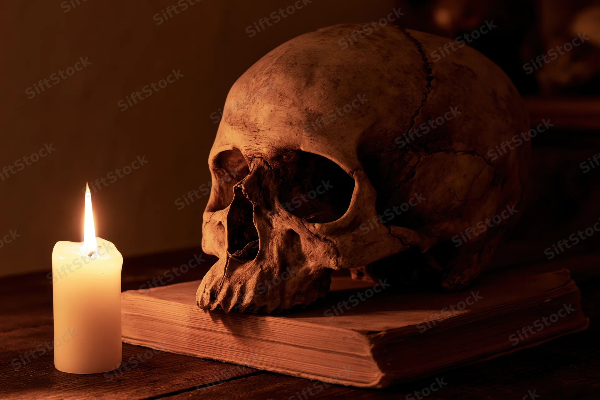 Human skull resting on an old worn book beside a lit candle on a wooden table