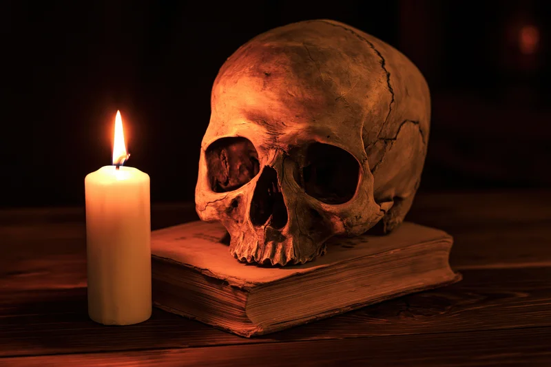 Human skull resting on an old worn book beside a lit candle on a wooden table