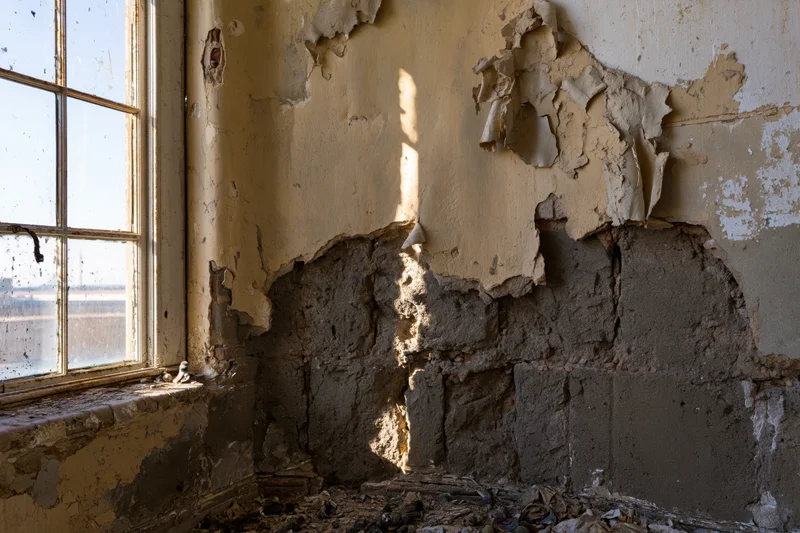 Crumbling plaster wall in an abandoned building corner near a window with sunlight and shadow