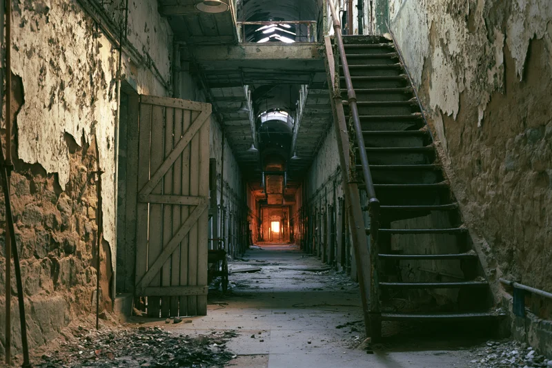Long abandoned prison corridor with crumbling walls and staircase leading into dim light at far end