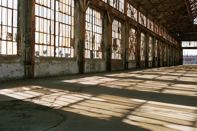 Wide view of derelict industrial warehouse interior with sunlight streaming through large windows casting grid shadows on cracked concrete floor