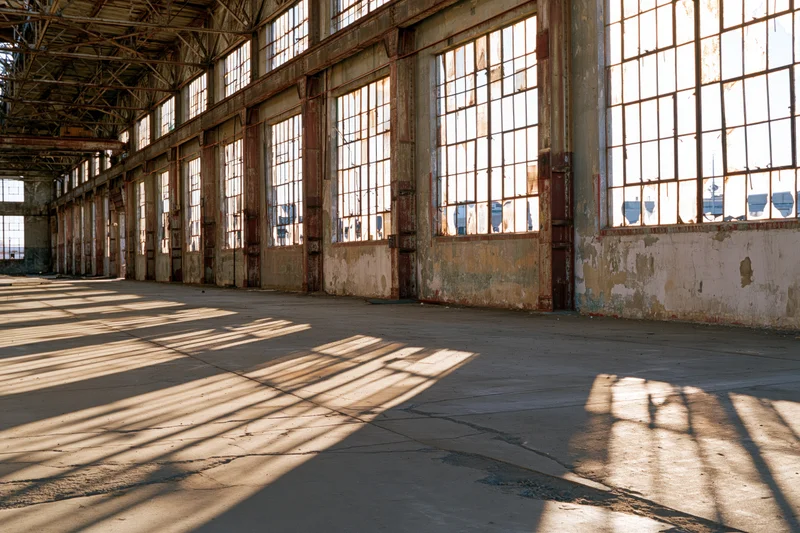 Wide interior view of derelict industrial warehouse with sunlight streaming through large windows casting shadows on concrete floor