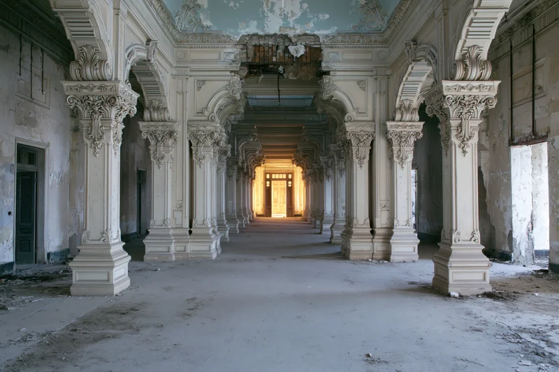 Long ornate hallway in abandoned colonial building with decaying columns and light at far end
