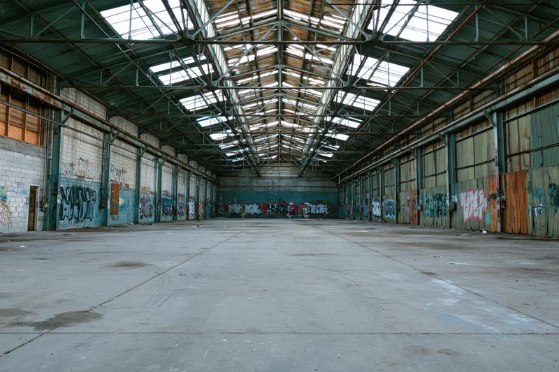Empty interior of a vast abandoned industrial warehouse with graffiti-covered walls and a high steel-framed ceiling