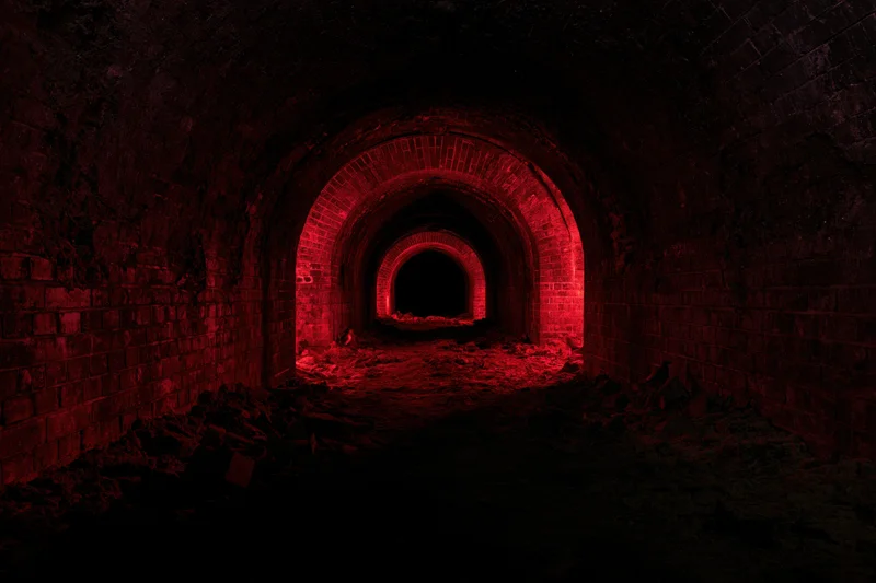 Dark red-lit arched brick tunnel with rubble inside abandoned underground cellar