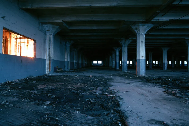 Wide interior view of an abandoned industrial warehouse with concrete pillars and debris on the floor