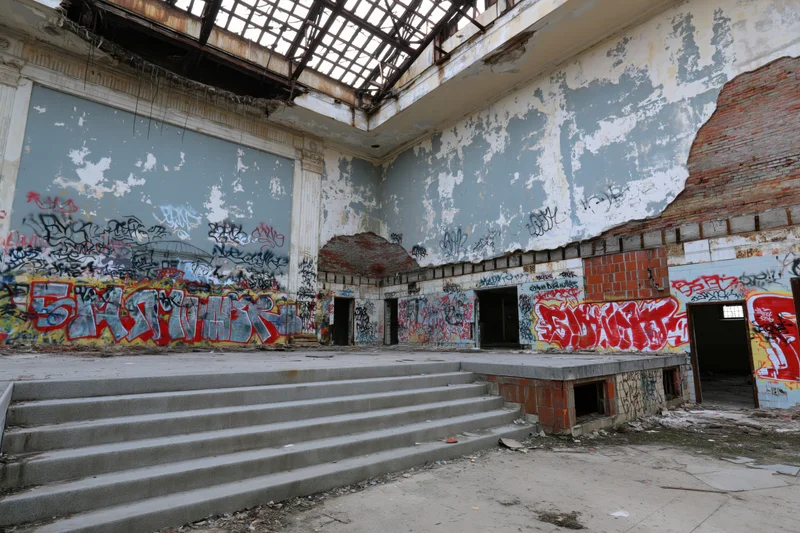 Abandoned hall interior with graffiti-covered walls and concrete stairs leading to a crumbling stage area