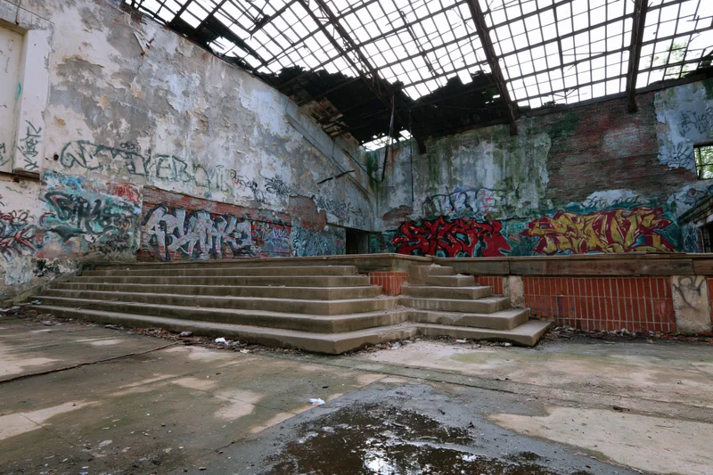 Abandoned hall interior with crumbling plaster walls and graffiti-covered concrete stairs under broken skylight roof