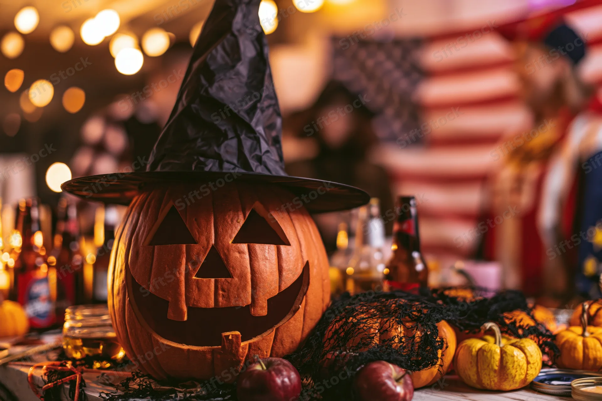 Carved pumpkin wearing witch hat on table at halloween party with drinks and decorations