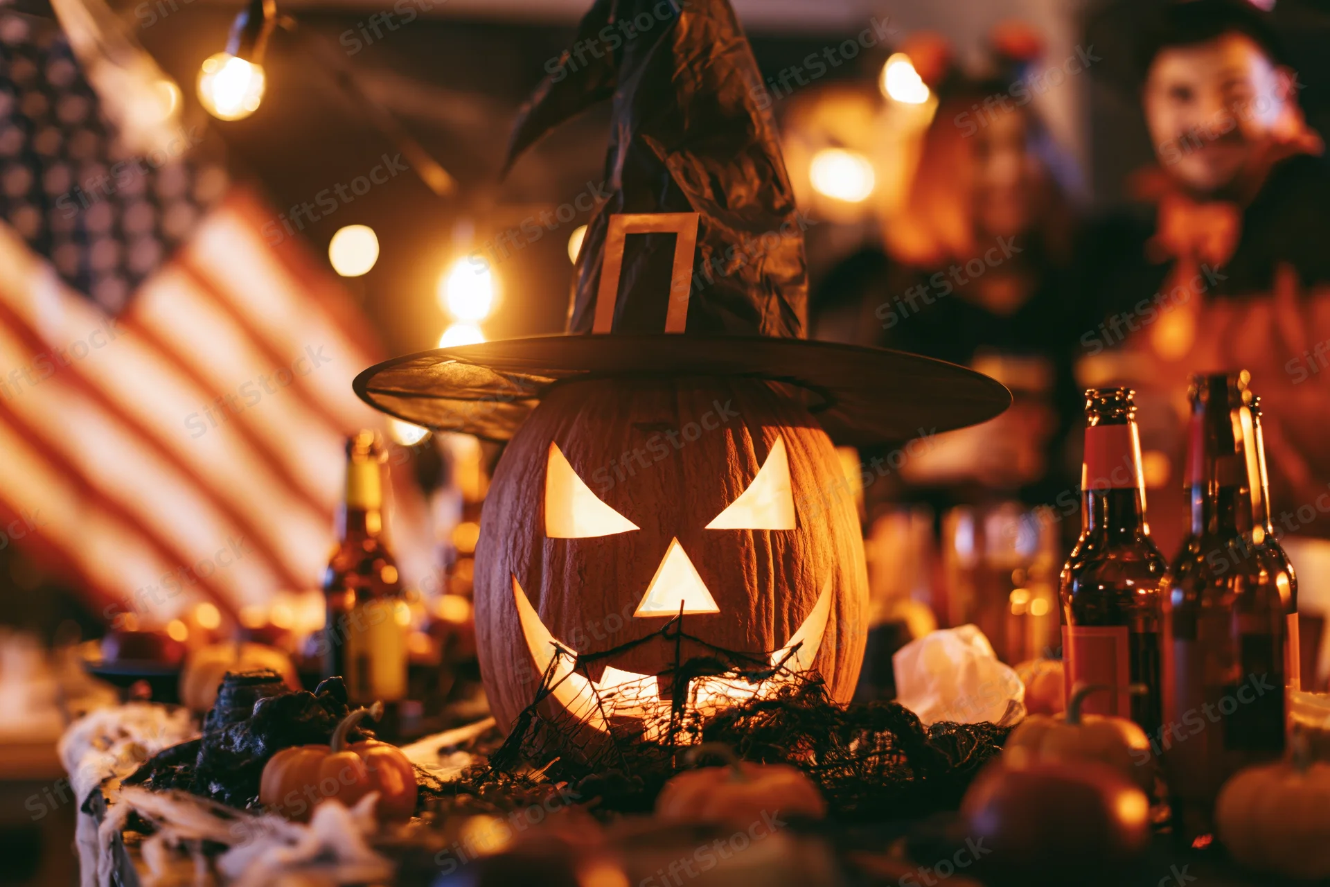 Carved jack-o-lantern wearing witch hat on table at halloween party with beer bottles and string lights