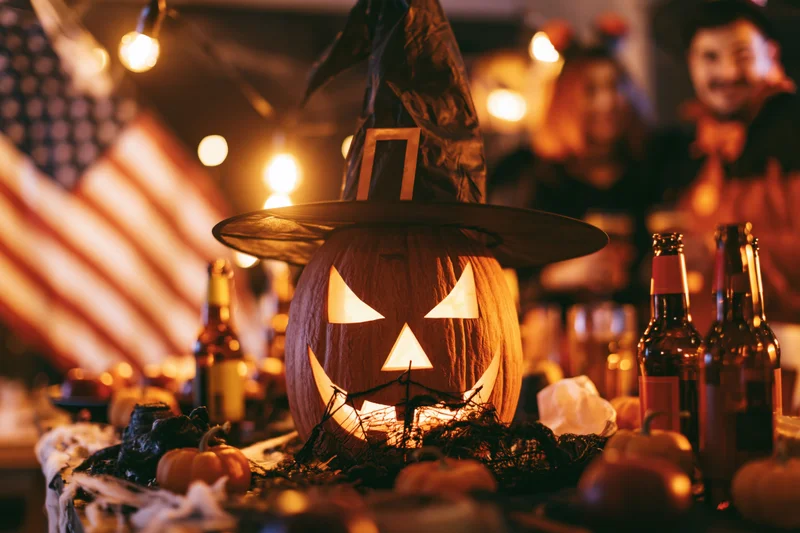 Carved jack-o-lantern wearing witch hat on table at halloween party with beer bottles and string lights