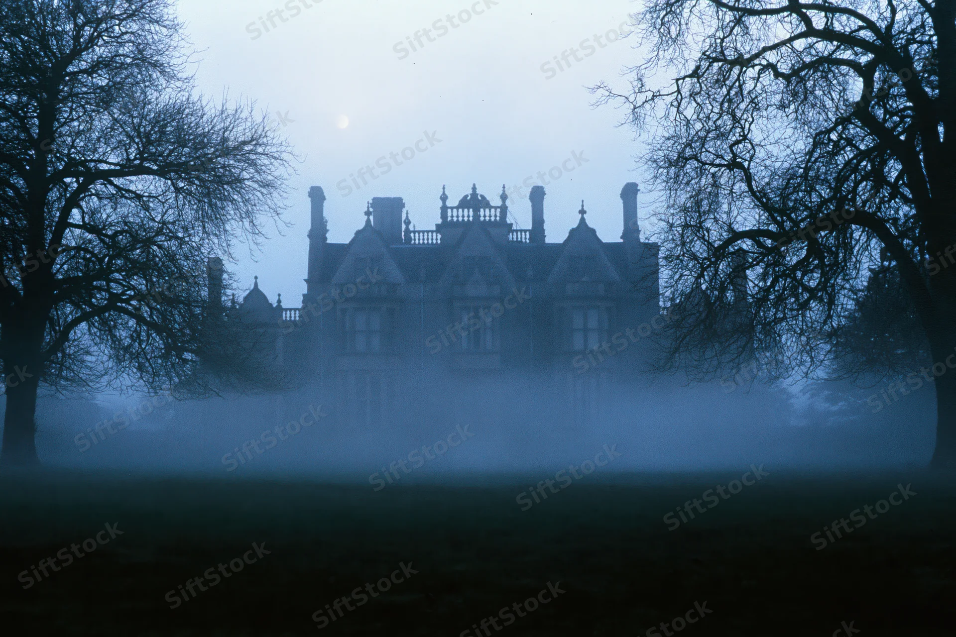 Victorian manor house silhouetted in dense fog at night surrounded by bare trees