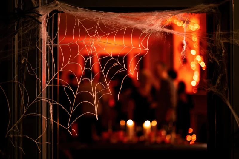 Decorative spiderweb stretched across a dark doorframe framing a dimly lit party scene with candles and orange lights