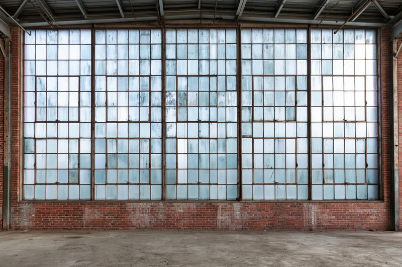 Large industrial warehouse interior with tall multi-paned windows set in brick wall, concrete floor, metal ceiling beams, empty space