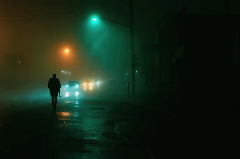 Silhouette of a man walking alone on a wet urban street at night under fog and streetlights