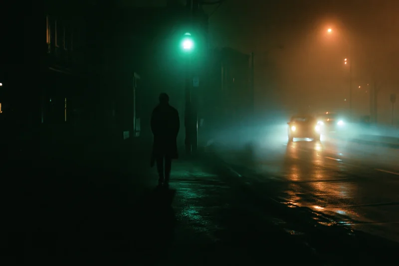 Lone figure walking along a wet urban street at night under green traffic light with car headlights in fog