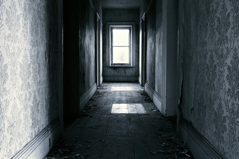 Long abandoned hallway with peeling wallpaper and debris leading to a single window at the end