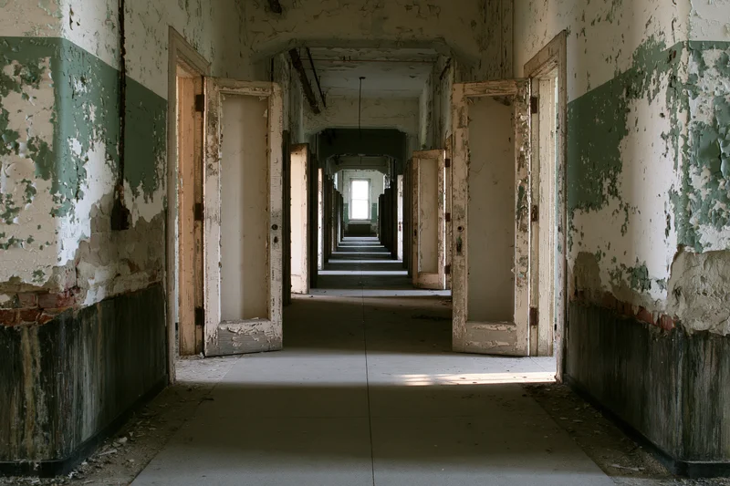 Long abandoned institutional hallway with peeling paint and open doors receding into distance