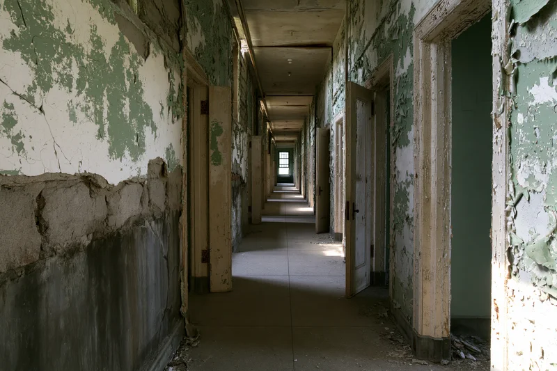 Long abandoned institutional hallway with peeling paint and broken plaster receding into the distance