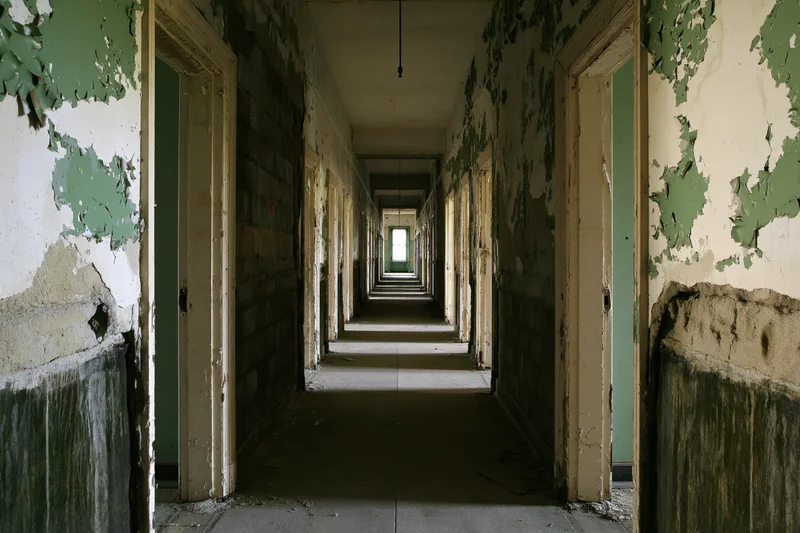 Long abandoned institutional hallway receding into the distance with peeling green paint and doors on either side