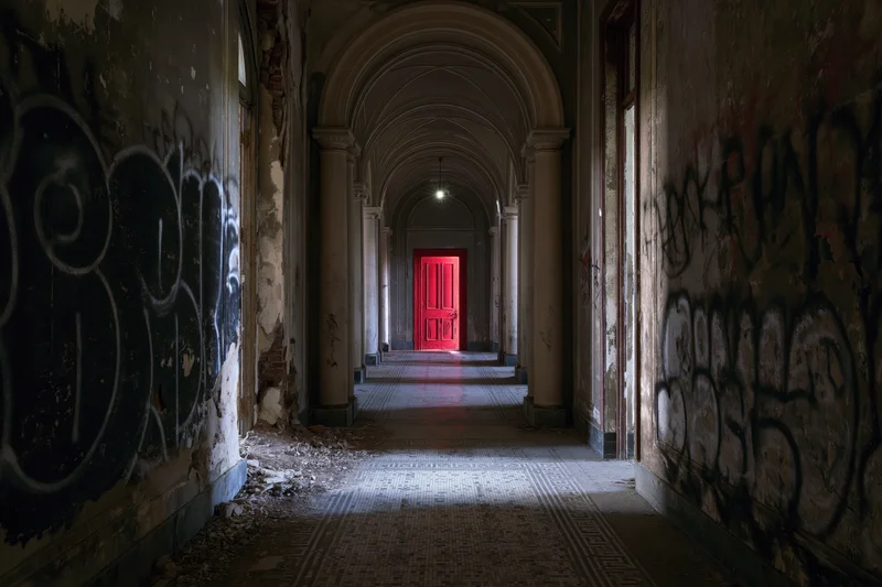 Long decaying corridor with red door at end, flanked by graffiti-covered walls and arched columns, tiled floor littered with debris