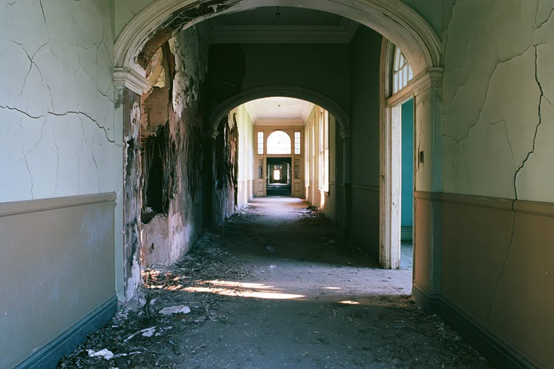Long decaying hallway in an abandoned institutional building with crumbling walls and debris on the floor