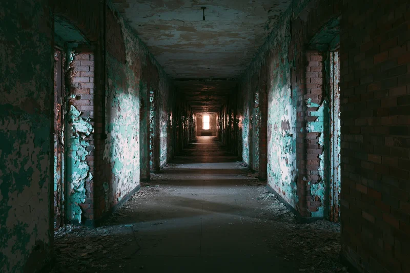 Long hallway in abandoned institutional building with peeling teal paint and exposed brick walls leading to distant light at end