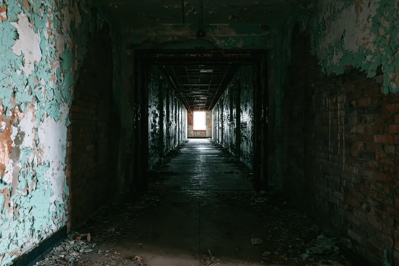 Long hallway in abandoned institutional building with peeling paint and brick walls leading to light at end