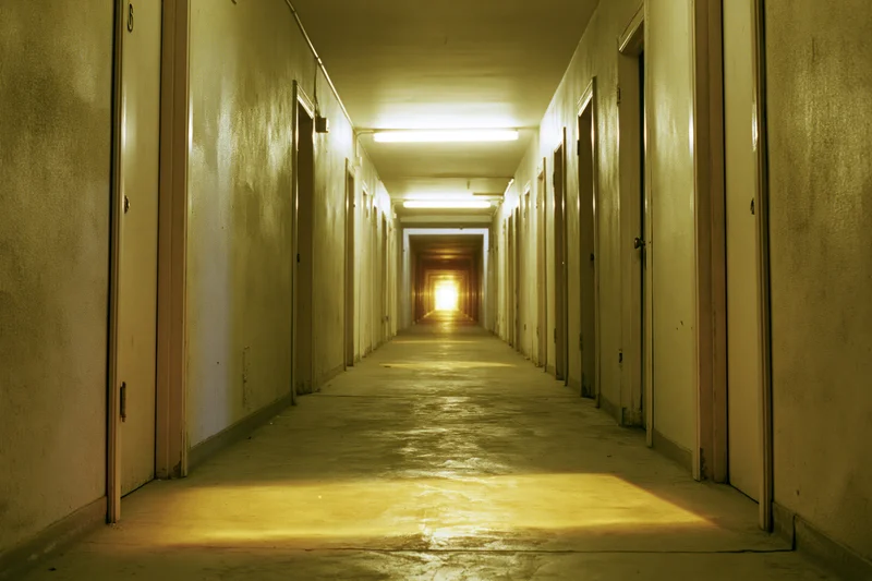 Long narrow institutional hallway receding toward a dim glow with fluorescent lights and doors on both sides