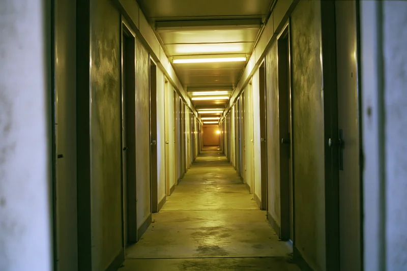 Long narrow institutional hallway receding toward a dim glow with fluorescent lighting and doors on both sides