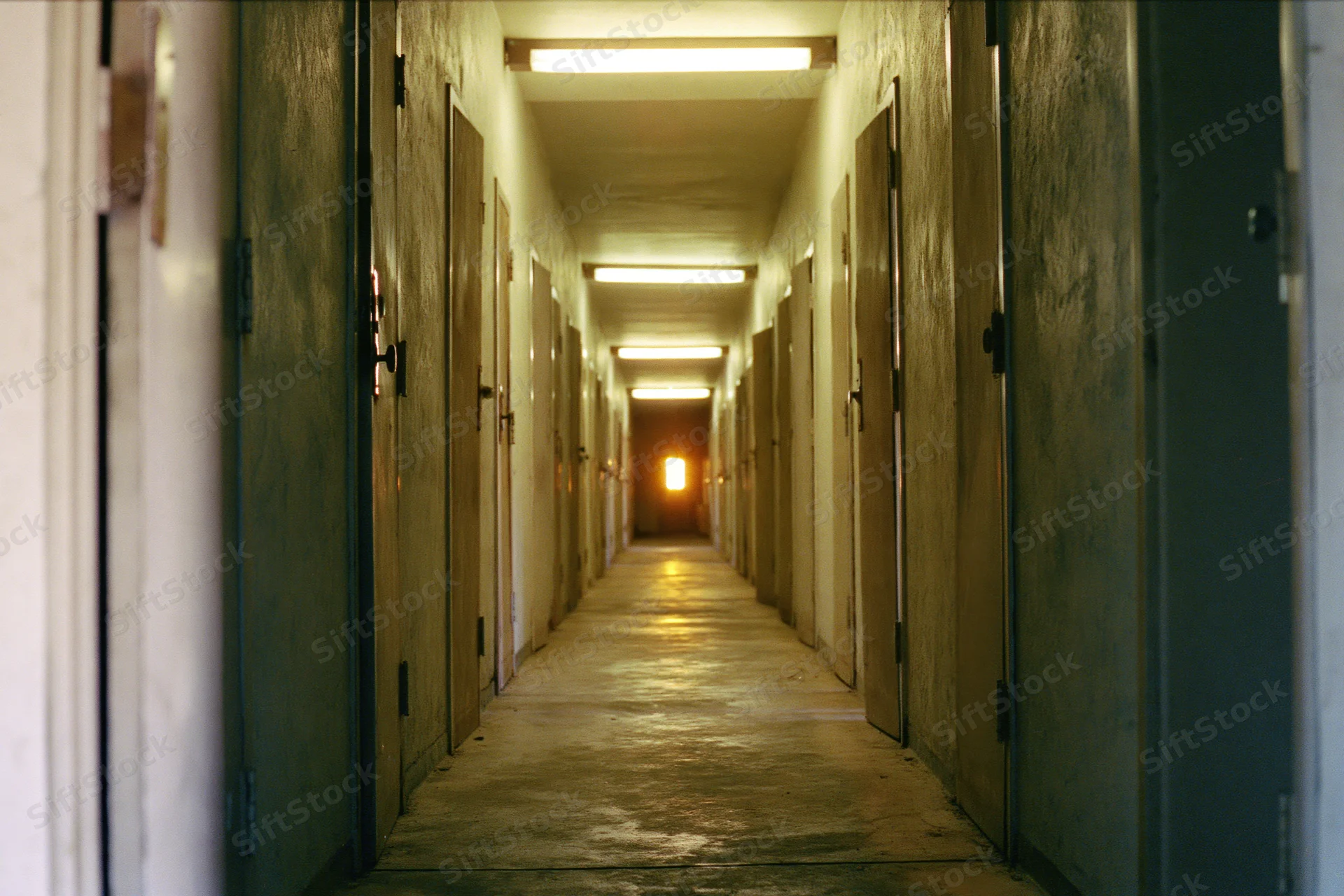 Long narrow institutional hallway receding toward a warm glowing end with doors on both sides