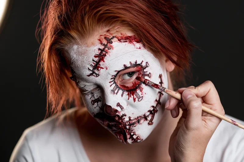 Woman applying detailed white and red face paint with brush around her eye for halloween horror costume