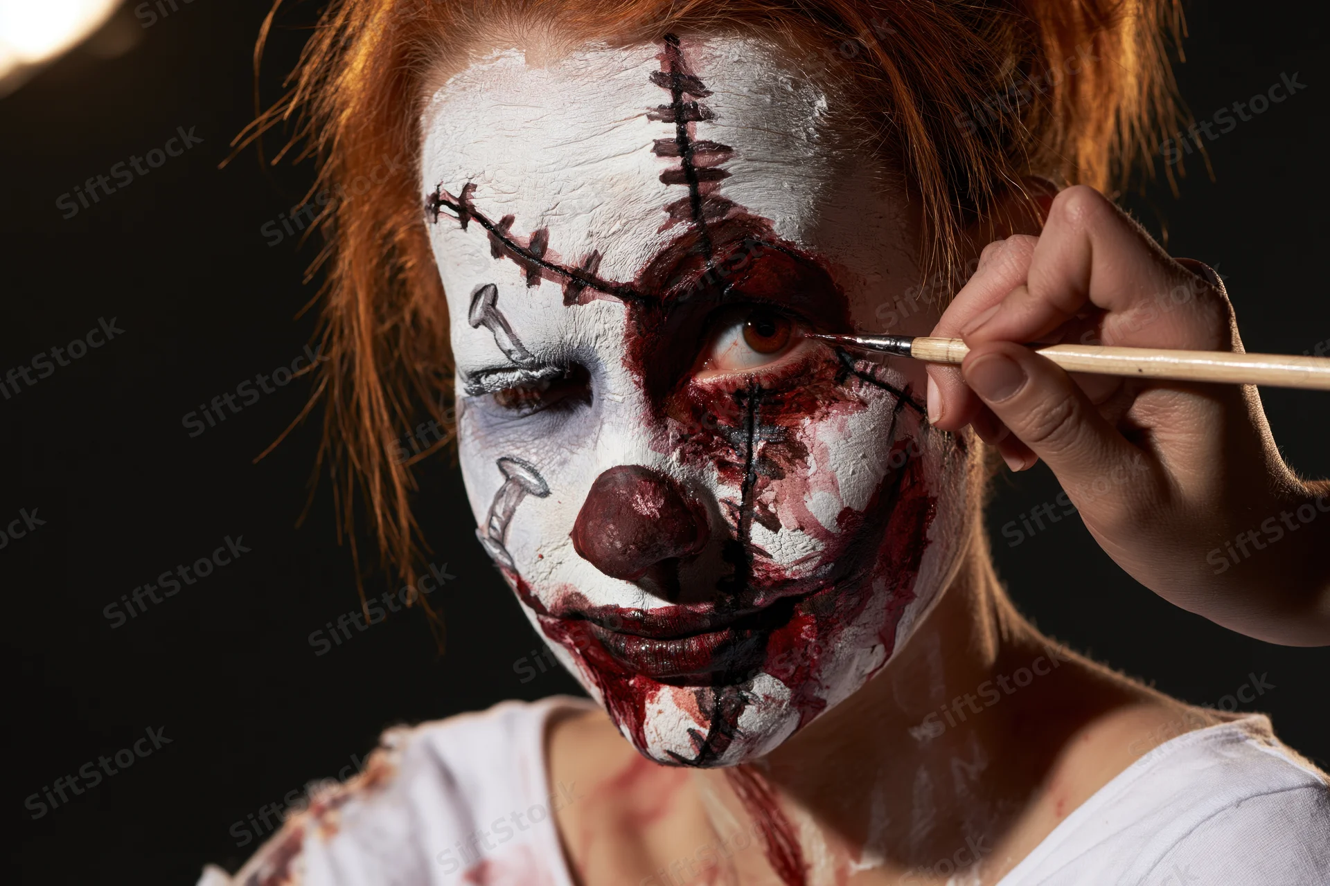 Woman applying detailed horror clown makeup with brush around eye in studio setting