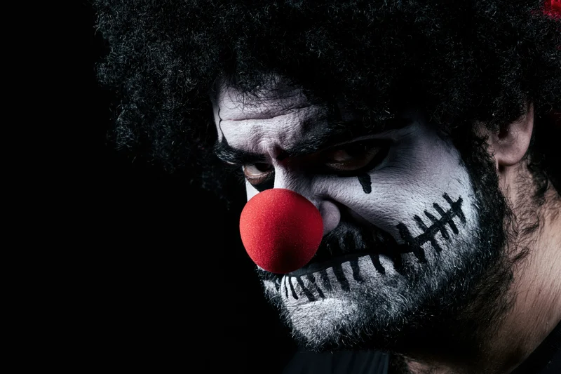 Closeup of a man with sinister clown makeup and red foam nose against dark background