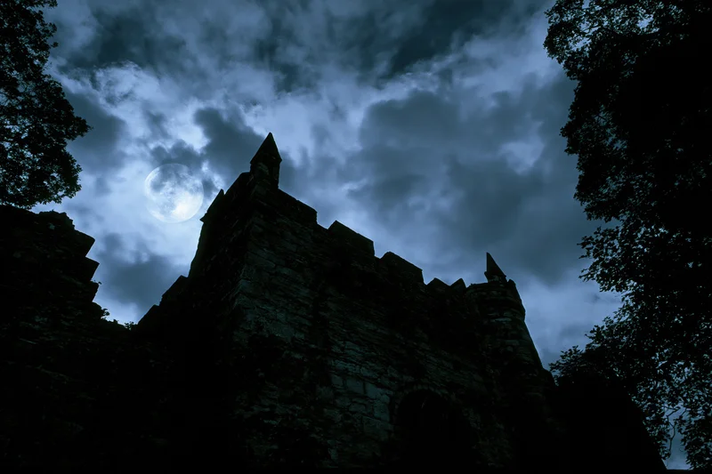 Medieval stone castle silhouette with pointed towers under moonlit cloudy sky at night