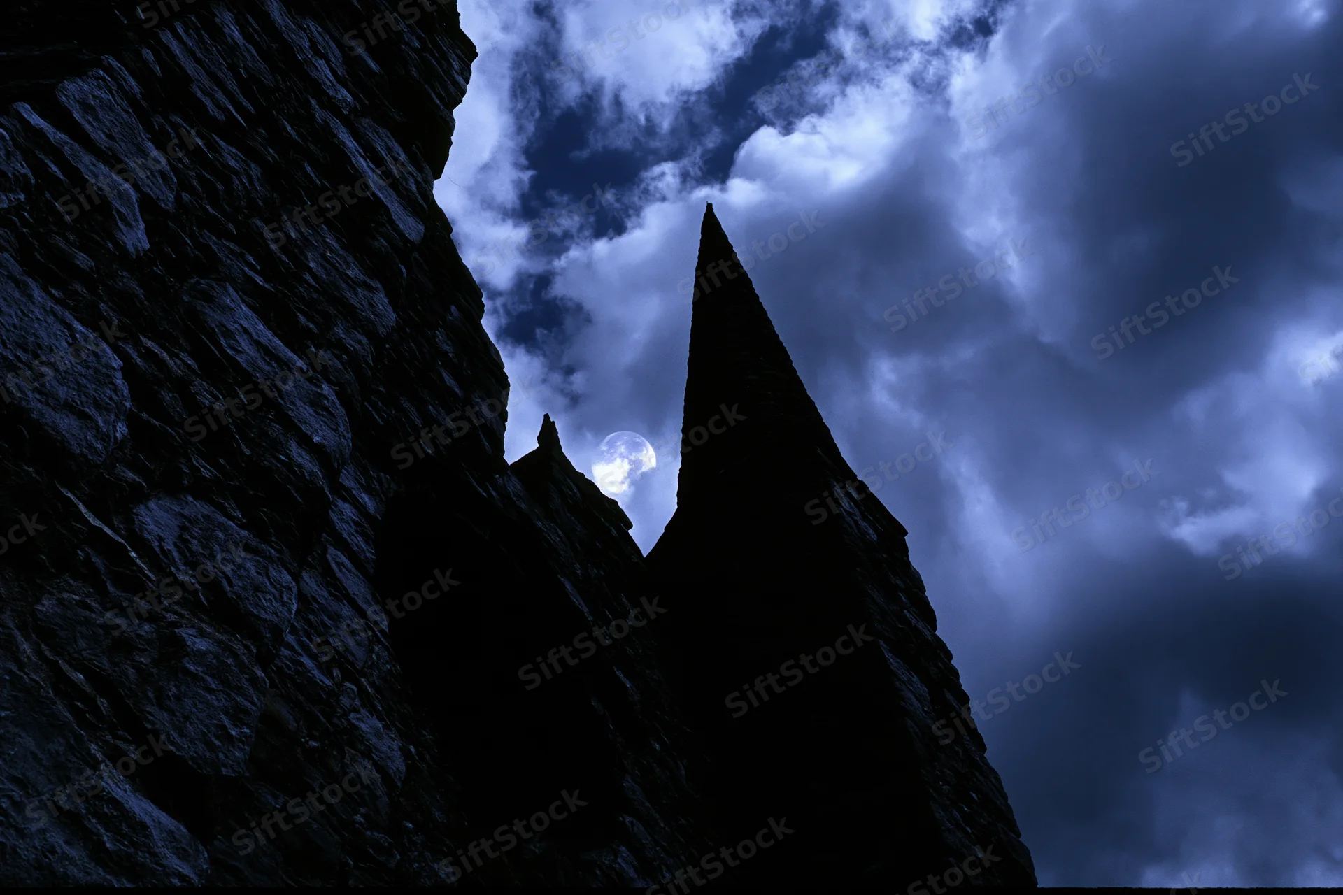 Medieval stone castle with pointed towers silhouetted against a cloudy night sky with moonlight