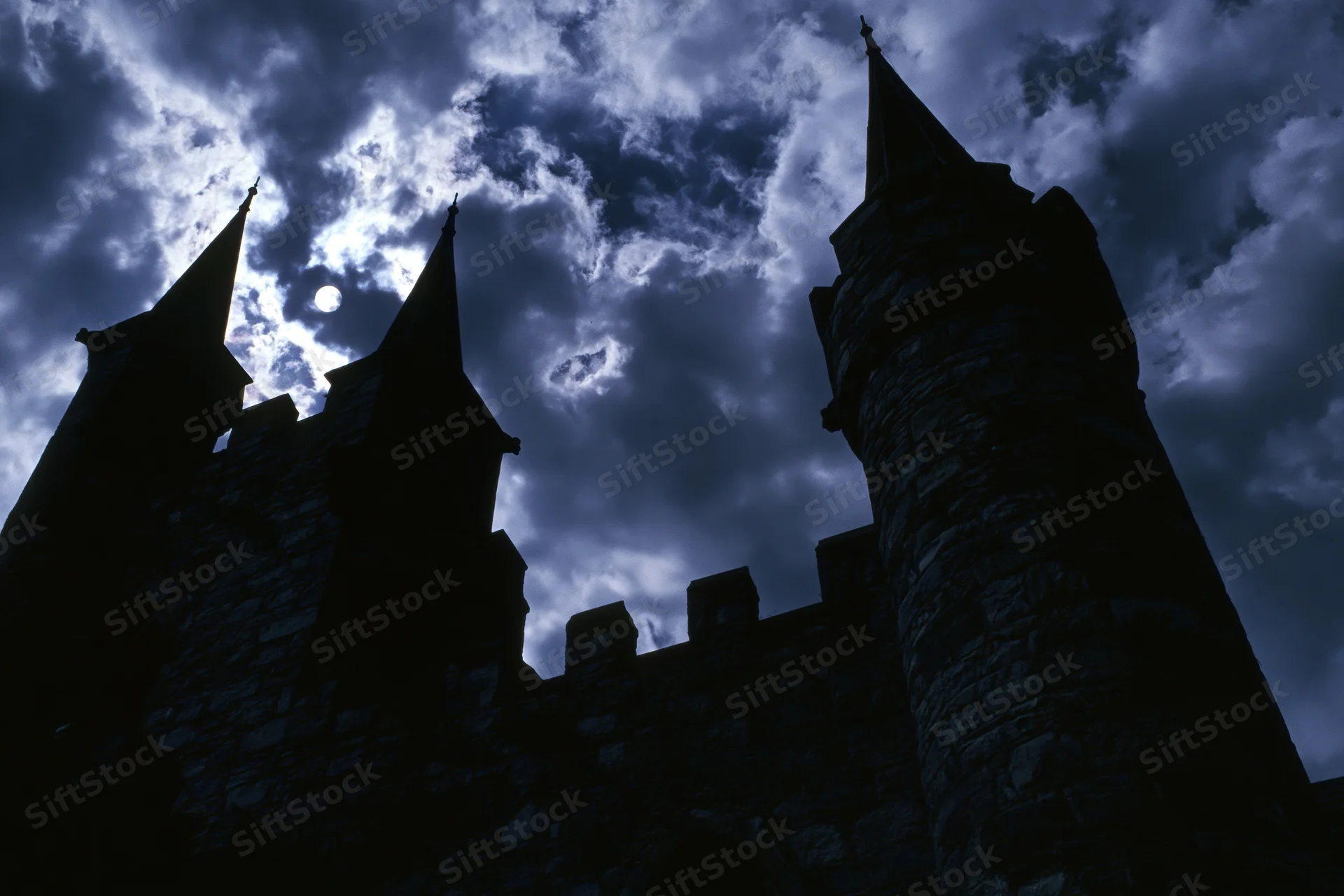 Medieval stone castle with pointed towers silhouetted against a cloudy night sky with moonlight