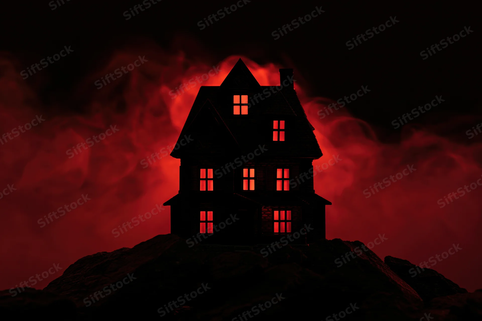 Miniature haunted house model glowing red from windows on rocky base against dark smoky background