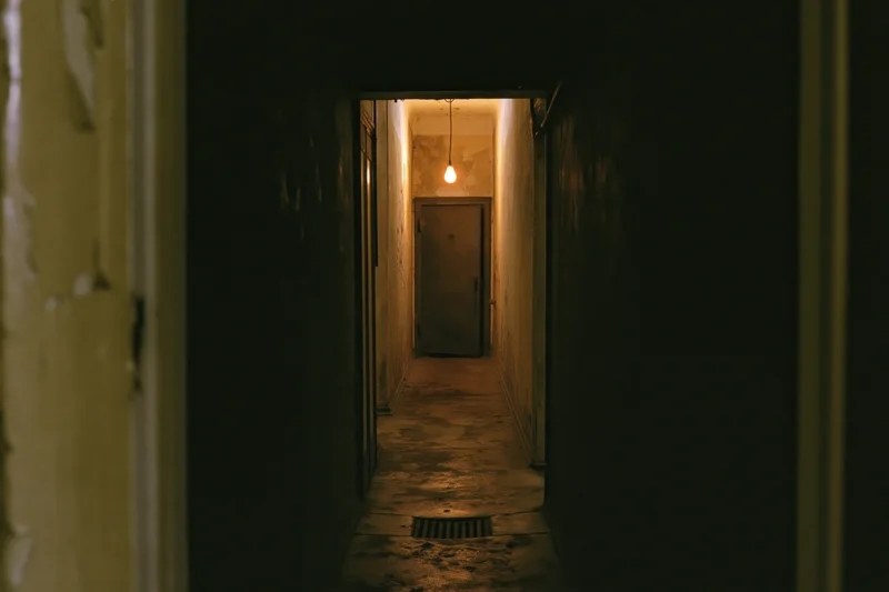 Narrow abandoned hallway receding toward a single metal door under dim warm light