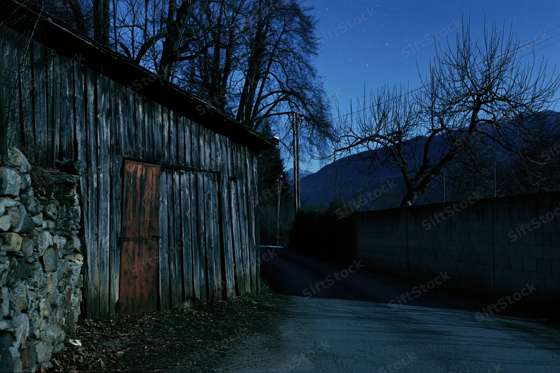 Narrow rural lane at night beside an old weathered wooden shed with bare trees and distant mountains under a starry sky