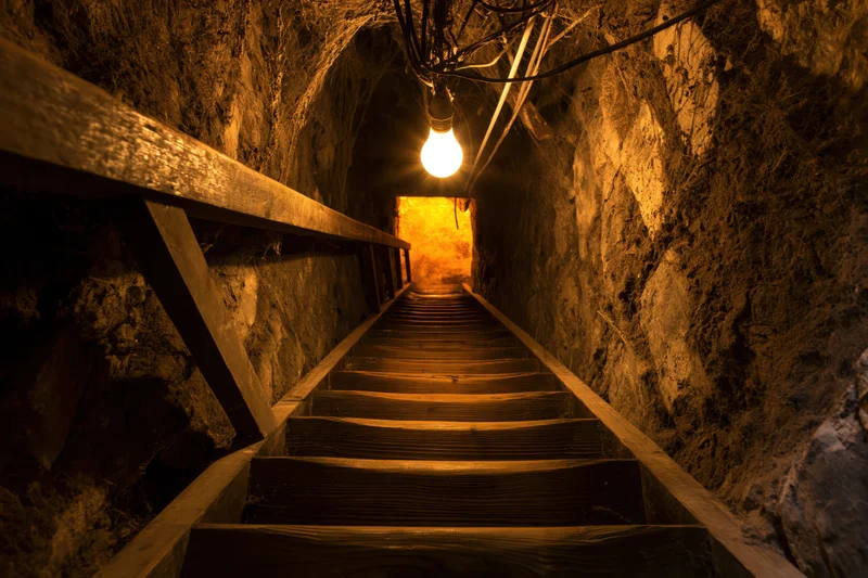 Wooden staircase descending into a narrow stone tunnel lit by a single bulb