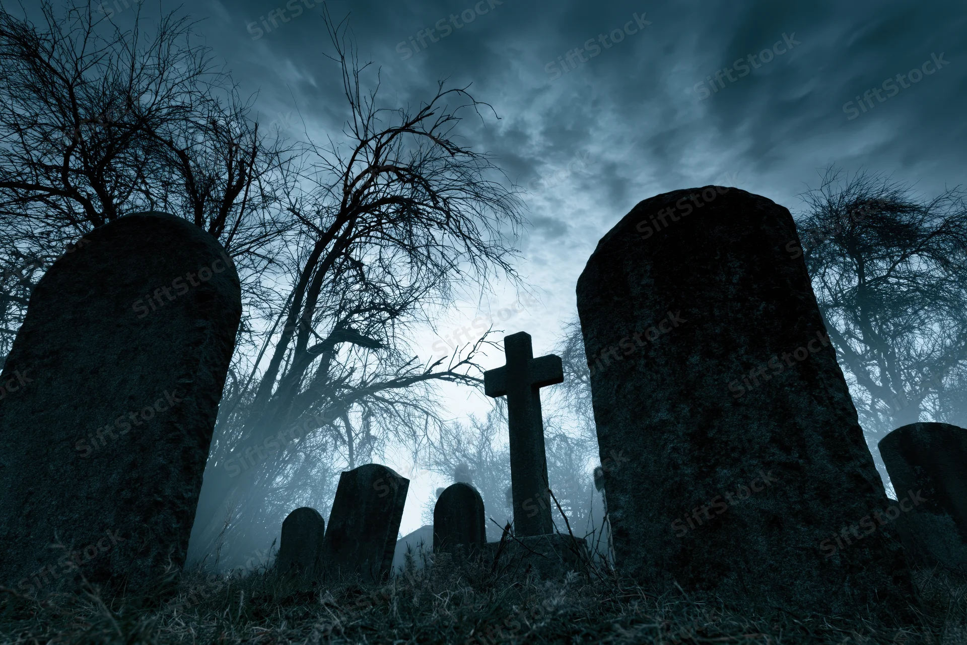 Weathered gravestones and a stone cross in a foggy graveyard at night