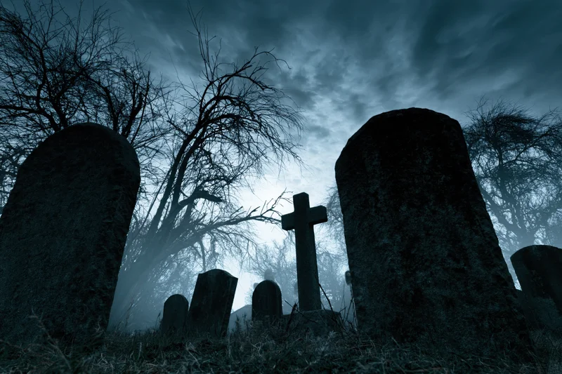 Weathered gravestones and a stone cross in a foggy graveyard at night