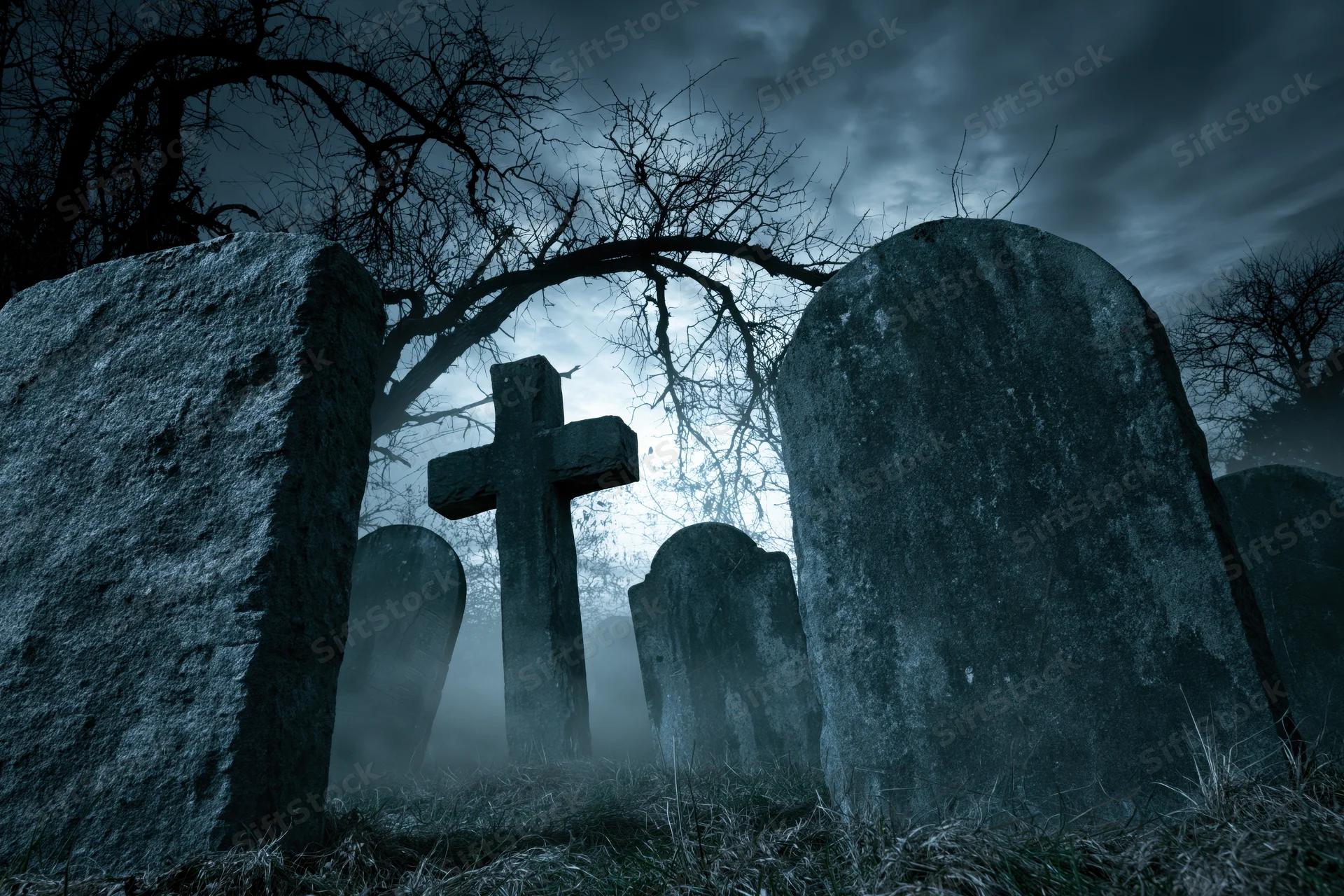Weathered gravestones and a stone cross in a foggy graveyard at night