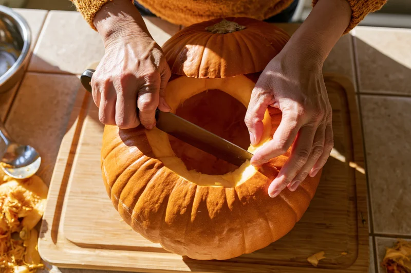 Hands carving a pumpkin on a wooden cutting board in a kitchen