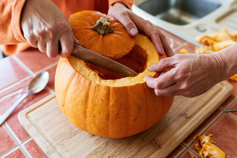 Hands carving a pumpkin on a wooden cutting board in a kitchen